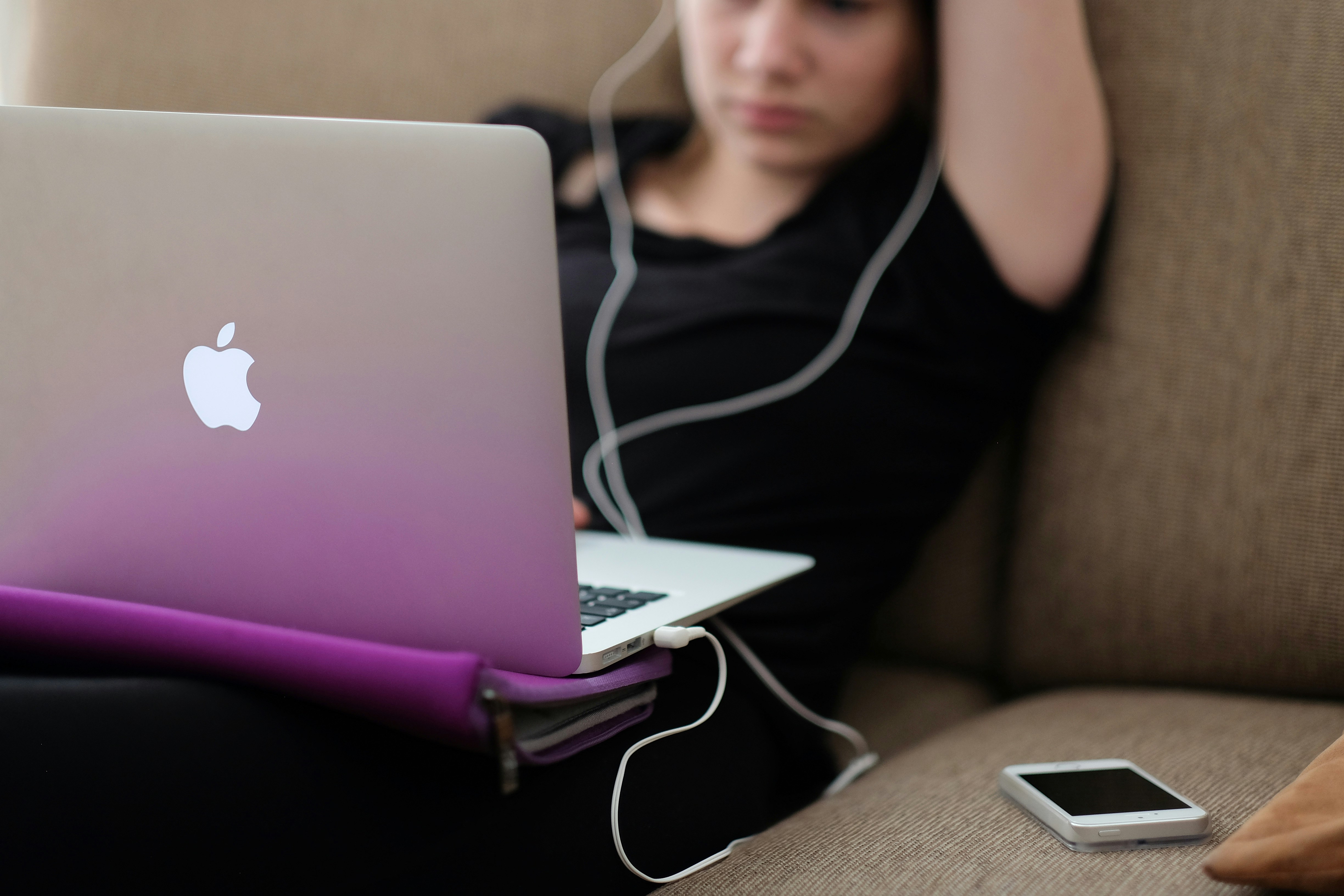 A woman using a laptop with earphones on a couch, with a smartphone nearby.